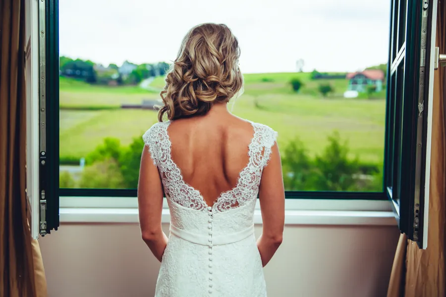 Braut von hinten im rückenfreien Spitzenkleid am offenen Fenster, draußen grüne Hügel