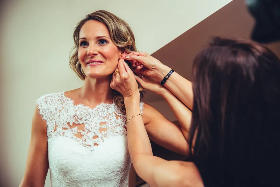 Braut im Spitzenkleid lächelt, während ihr eine Frau den Ohrring anlegt