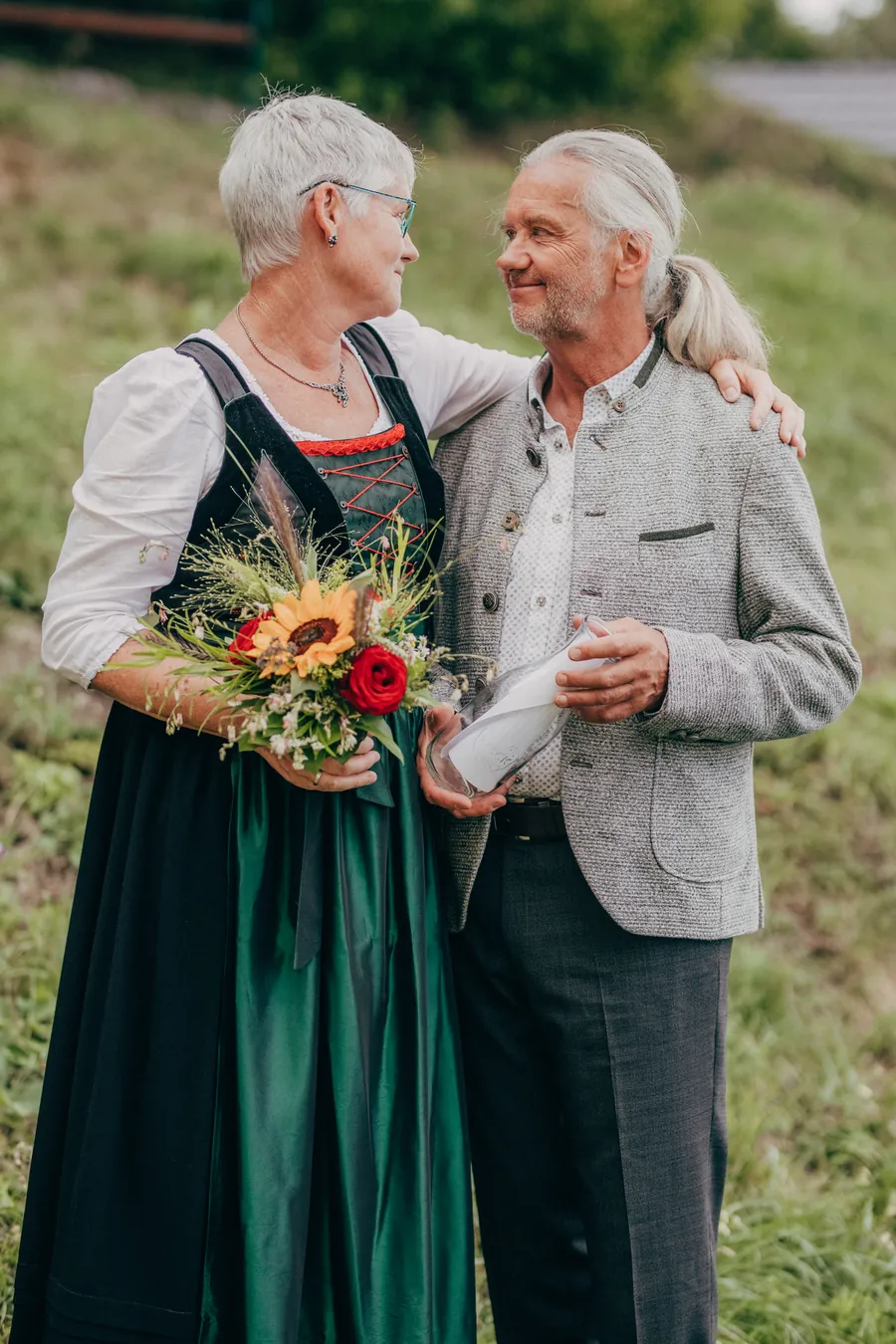 Brautpaar in Tracht Arm in Arm an einem grünen Hang, beide lachen sich an