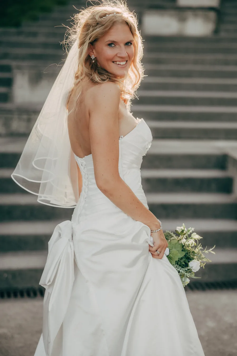 Braut steht auf einer Steintreppe und blickt lächelnd über die Schulter, rückenfreies Kleid mit Schleier