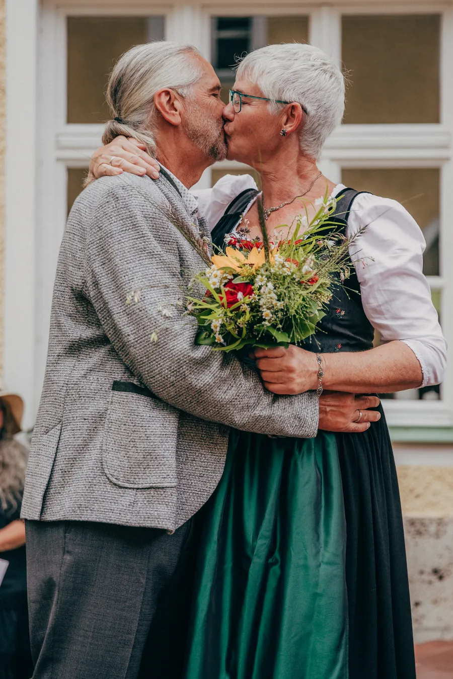 Brautpaar in Tracht küsst sich nach der Trauung vor einem Sprossenfenster, zwischen ihnen ein Wiesenblumenstrauß